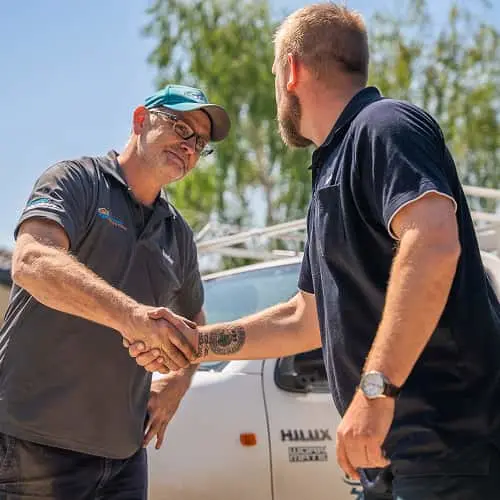 Two men in dark polo shirts shaking hands outdoors near a white vehicle with trees in the background.