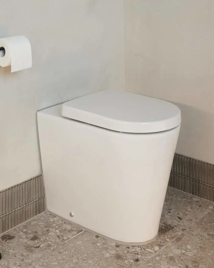 White modern toilet with closed lid in corner of bathroom with beige walls, gray speckled floor, and toilet paper roll.