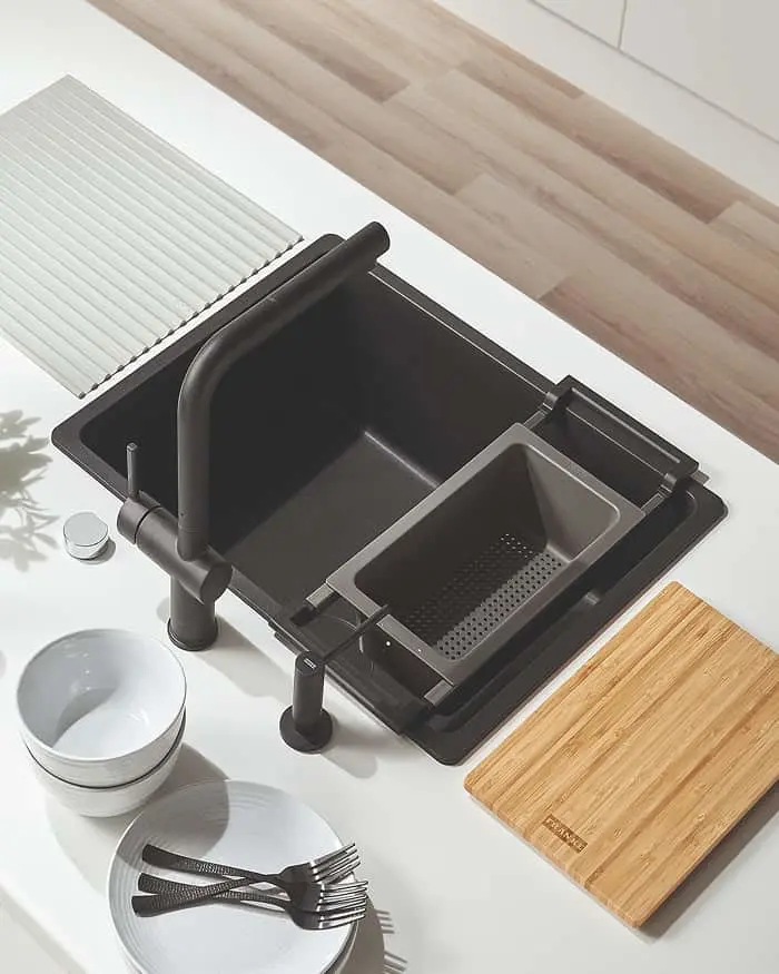 Black dish drying rack with two tiers and a bamboo cutting board on white countertop with dishes and utensils