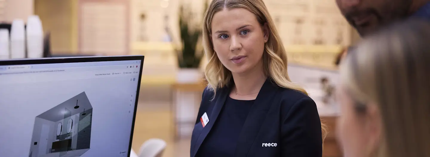 Woman in dark blazer with name tag standing next to computer monitor displaying architectural design in office setting.