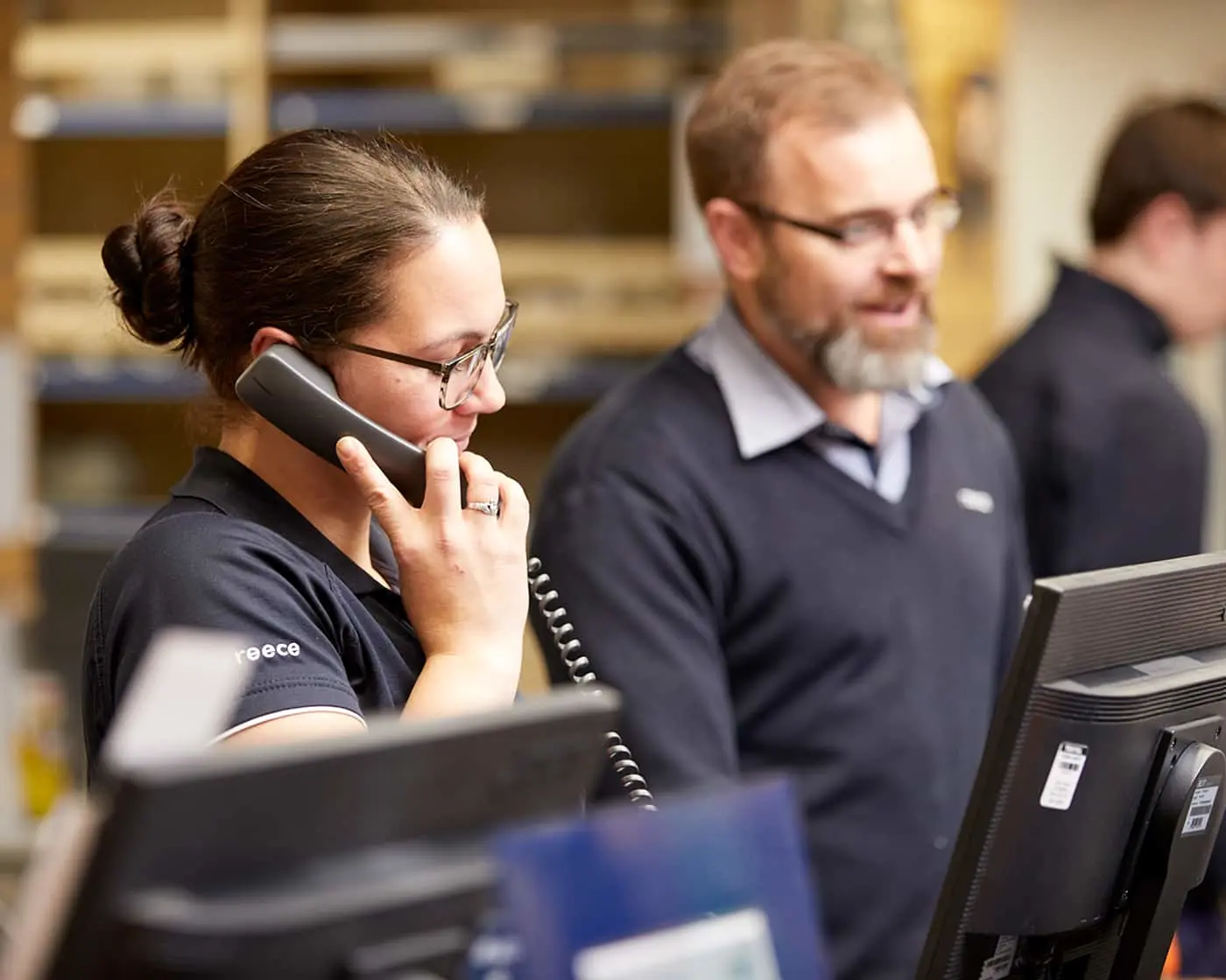 Woman with glasses talking on phone at desk with male colleague in background wearing dark polo shirts
