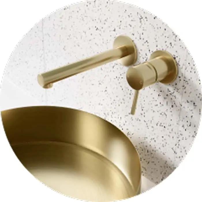 Brushed gold wall-mounted faucet above a gold sink basin against white terrazzo tile with black speckles.