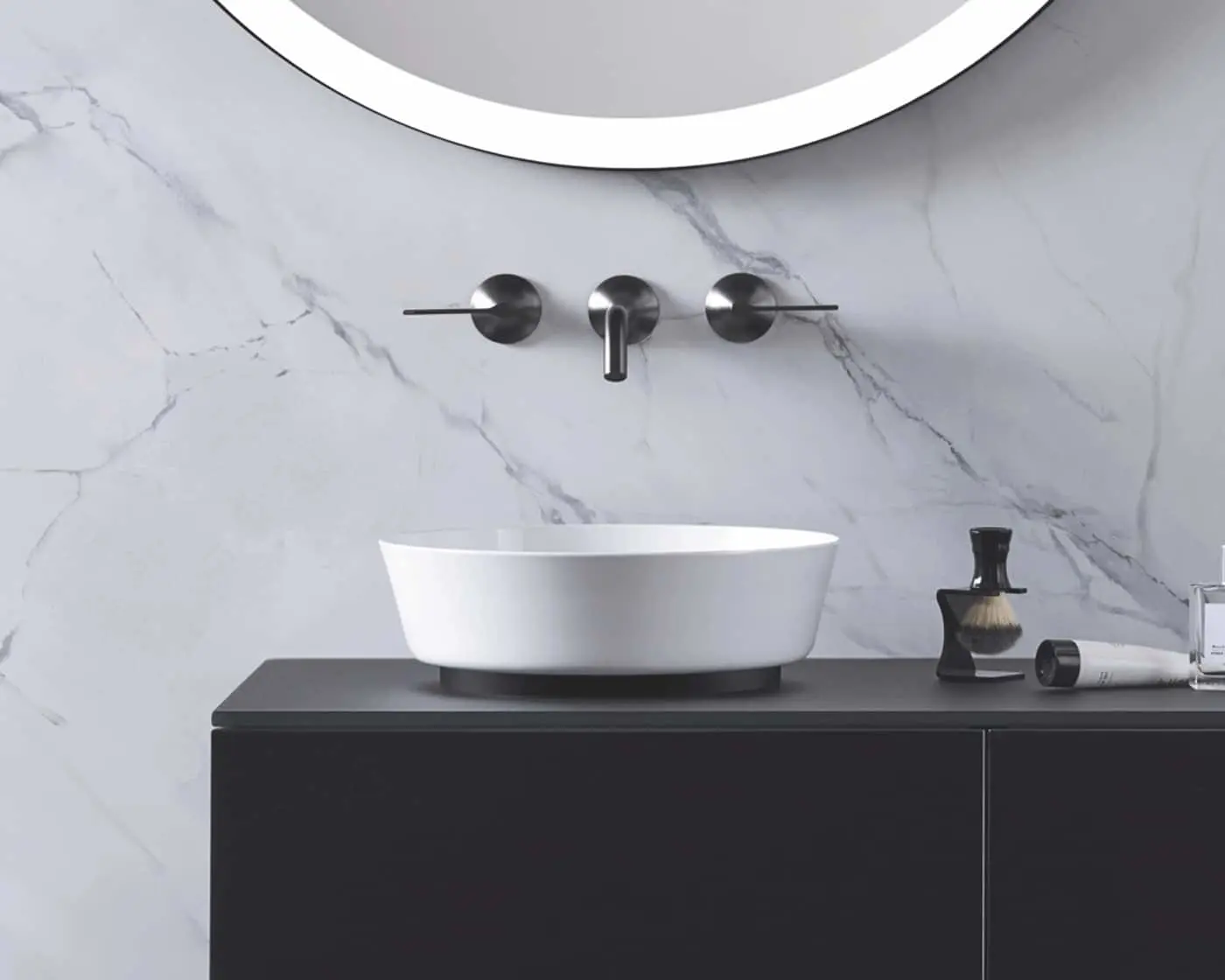 White vessel sink on dark vanity with wall-mounted faucet against marble backsplash and round mirror above.