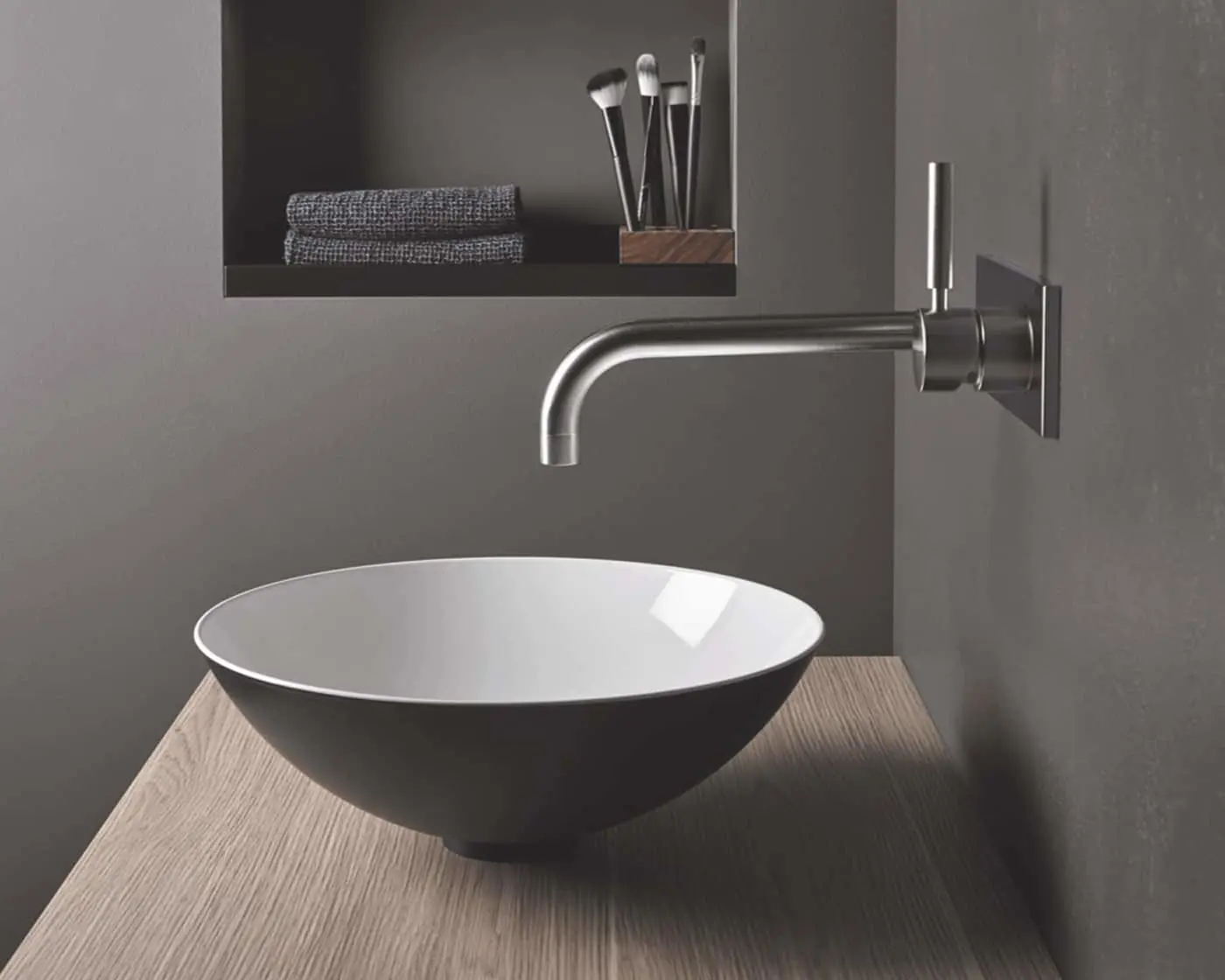 Modern bathroom with round white vessel sink on wood counter, wall-mounted faucet, and shelf with towels and brushes.