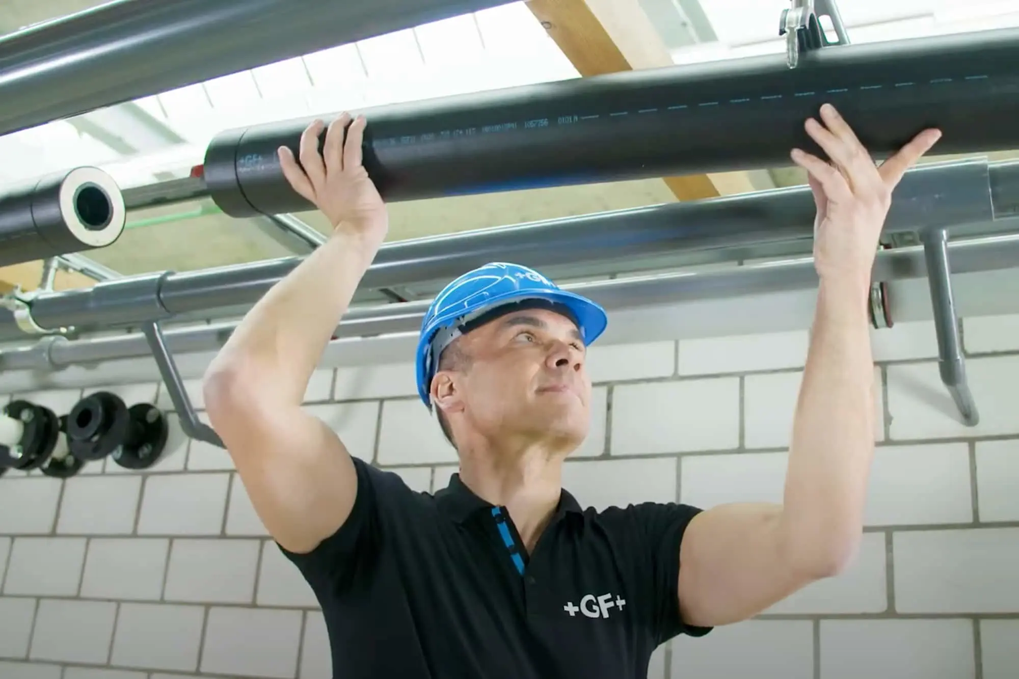 Worker in blue hard hat installing black pipe on ceiling with white tiled wall in background.