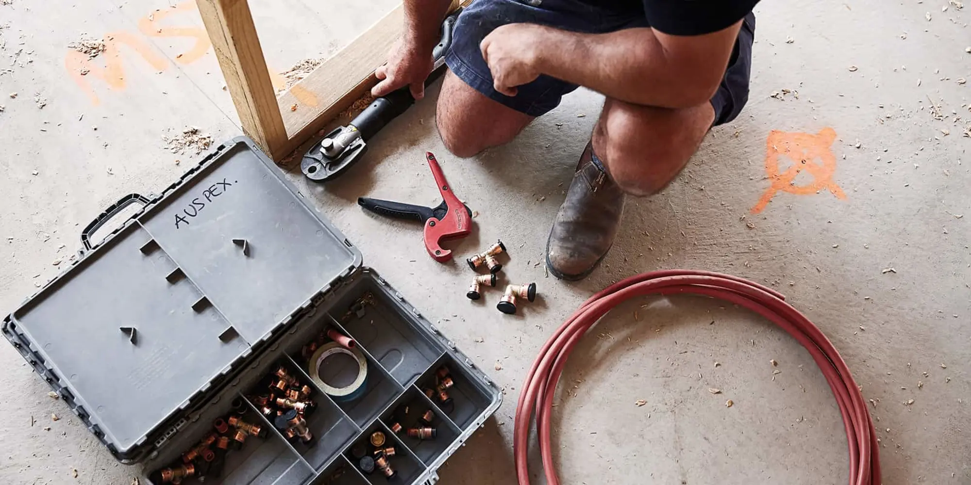 Person kneeling on concrete floor working with plumbing tools, toolbox with fittings, red pipe cutter, and coiled tubing.