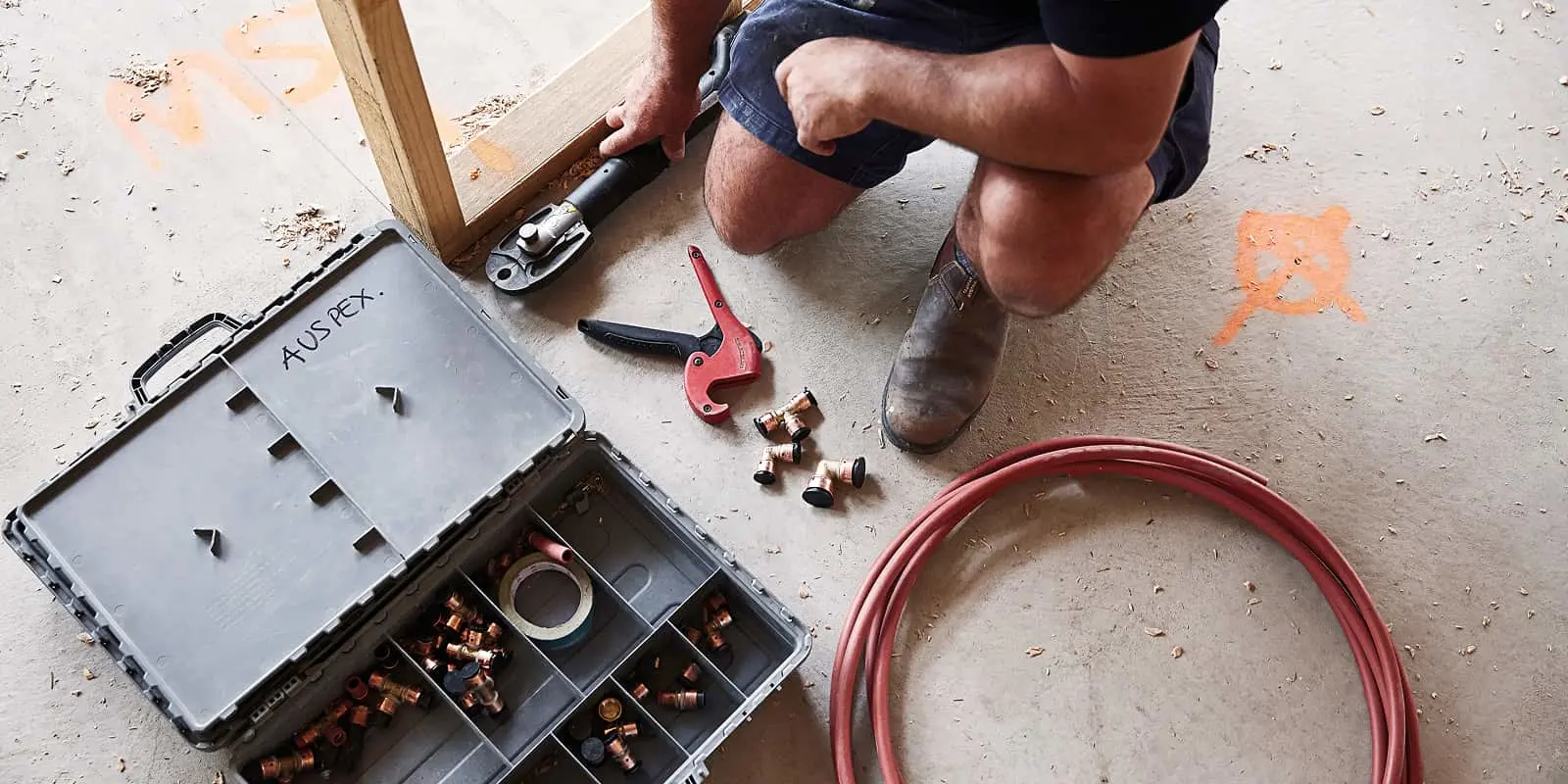 Person kneeling on concrete floor working with plumbing tools, toolbox with fittings, red pipe cutter, and coiled tubing.
