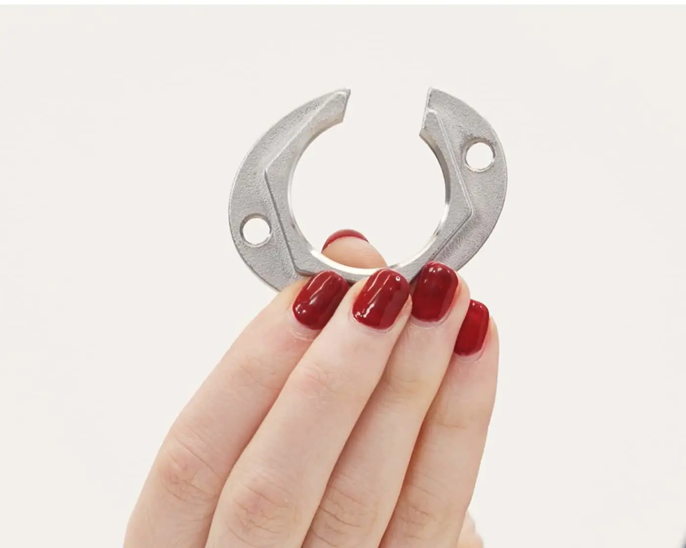 Hand with red nail polish holding a silver C-shaped metal clip with two mounting holes against a white background.