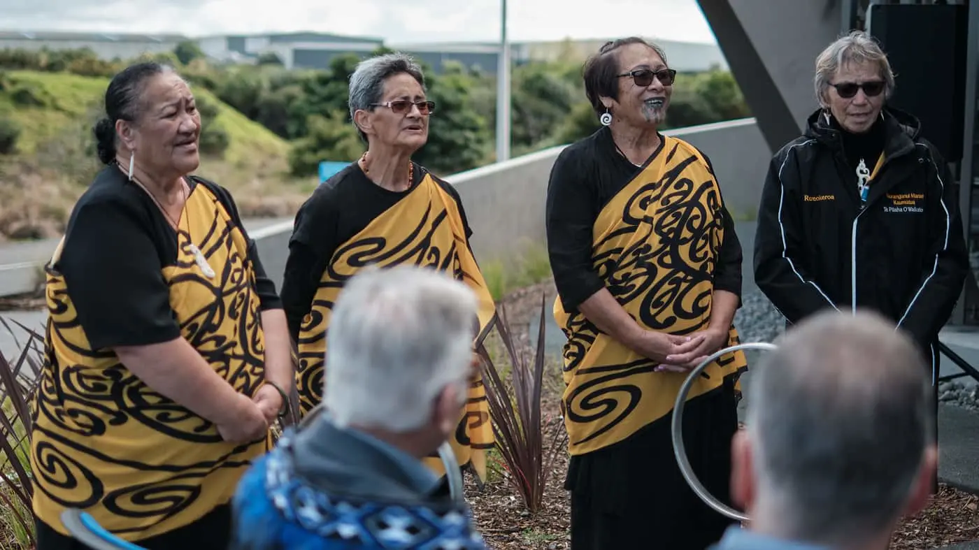 Four women in traditional yellow and black patterned garments stand outdoors addressing a seated audience.