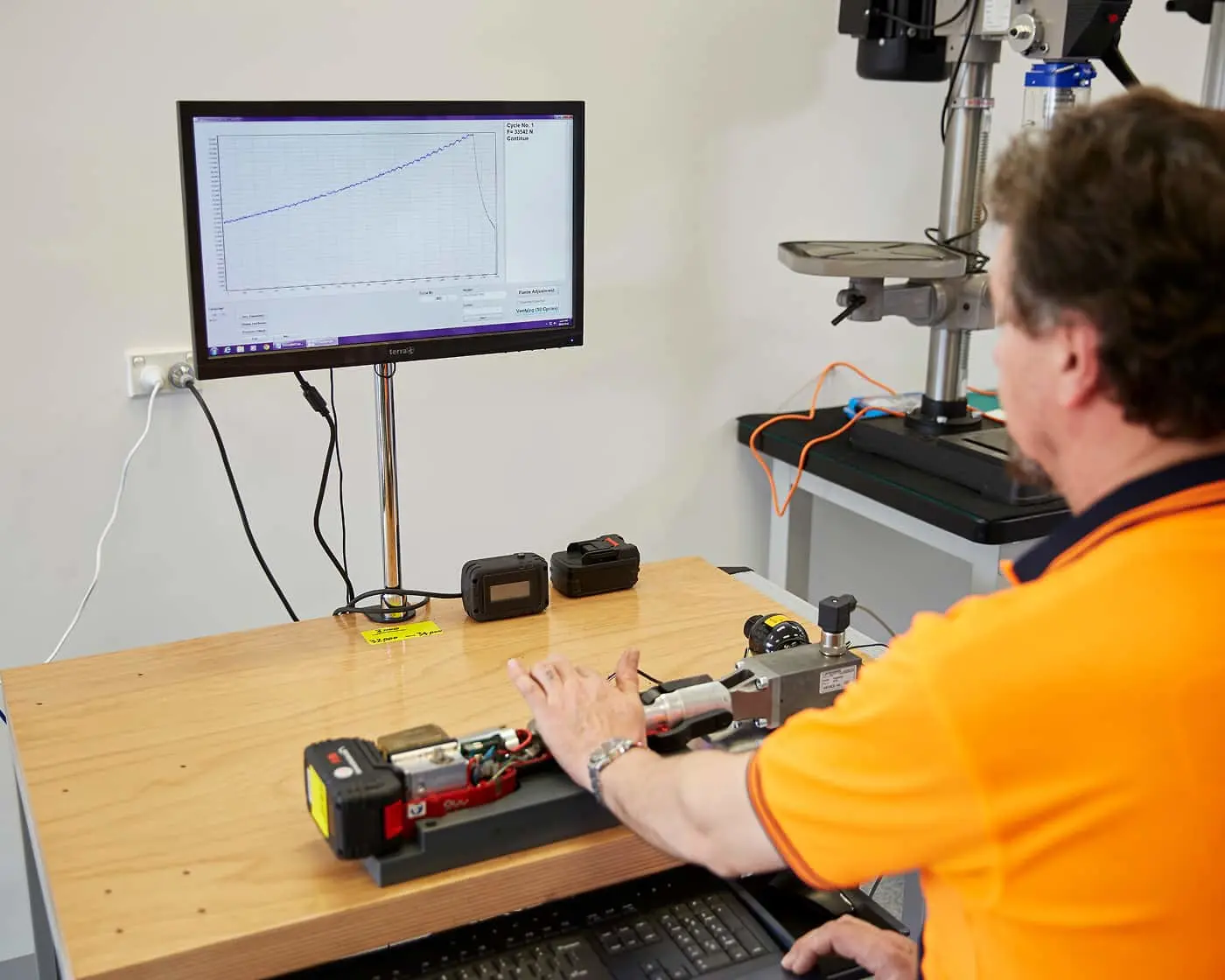 Person in orange safety vest viewing data graph on monitor while working with equipment on wooden desk.