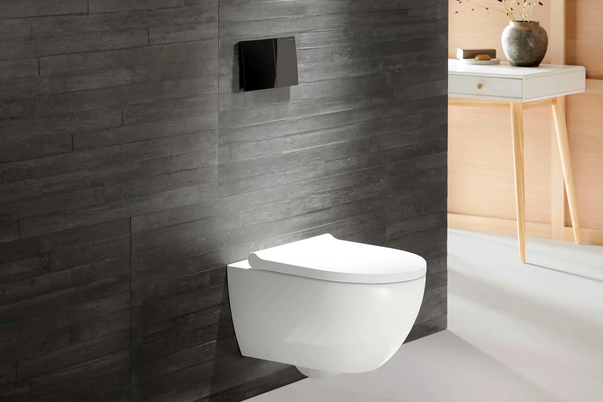 White wall-mounted toilet against dark gray tiled wall with black flush plate and white side table with vase.