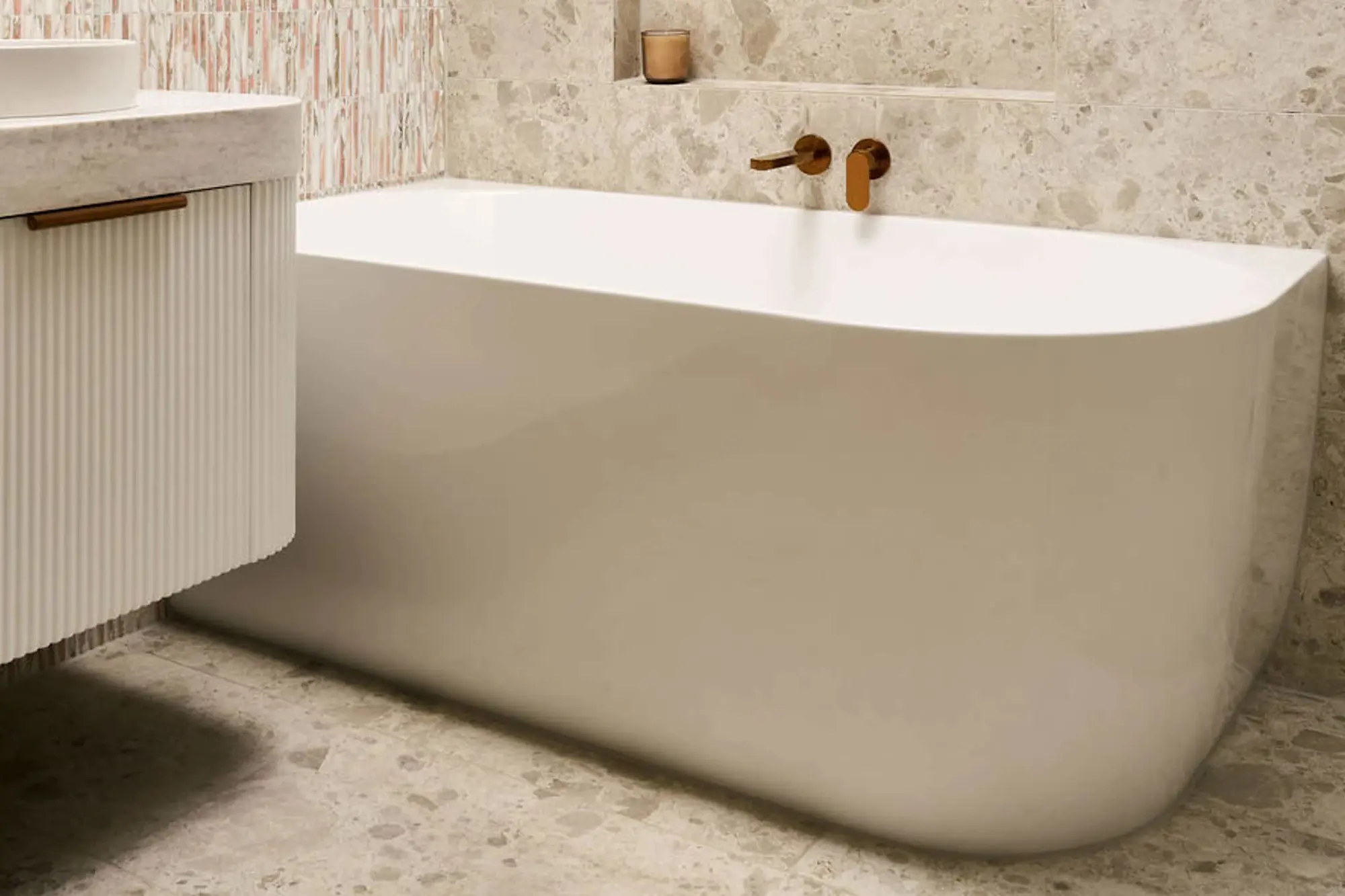 White freestanding bathtub with wall-mounted brass faucet against beige stone tile wall in modern bathroom.