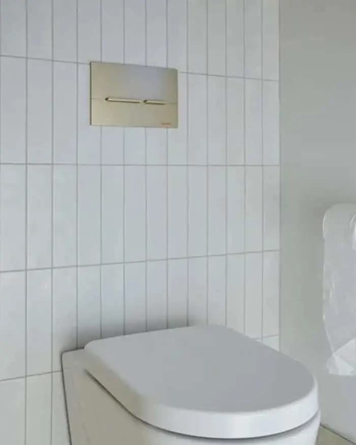 White toilet with closed lid against white tiled wall with rectangular flush plate mounted above