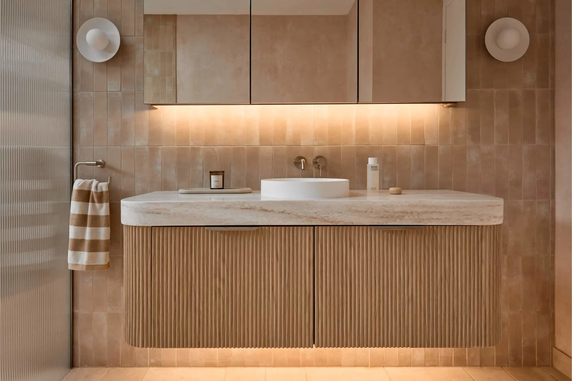 Modern bathroom vanity with white vessel sink, beige tile walls, under-cabinet lighting, and fluted wood cabinet.