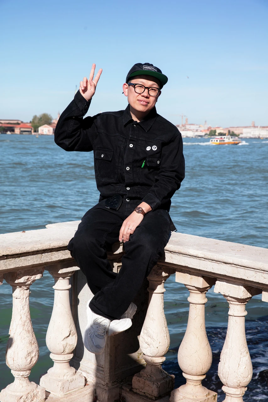 A young man in all-black attire and a cap sits on a stone balustrade, flashing a peace sign, with a scenic river and blue sky in the background.