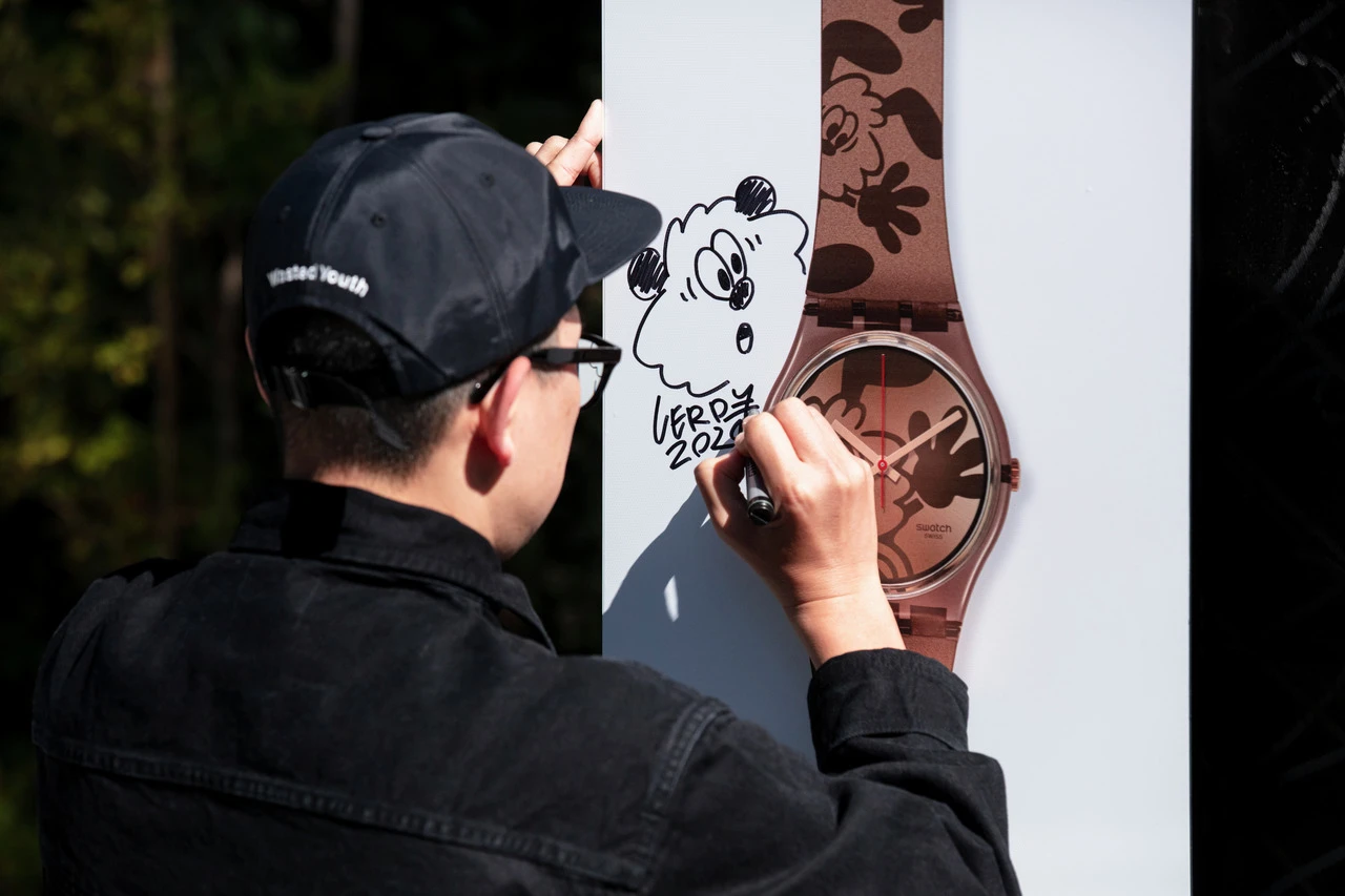 A person in a black jacket and cap is writing or drawing on a brown patterned column with a round, copper-toned object attached to it. there is a cartoon-like sheep drawing above their hand.