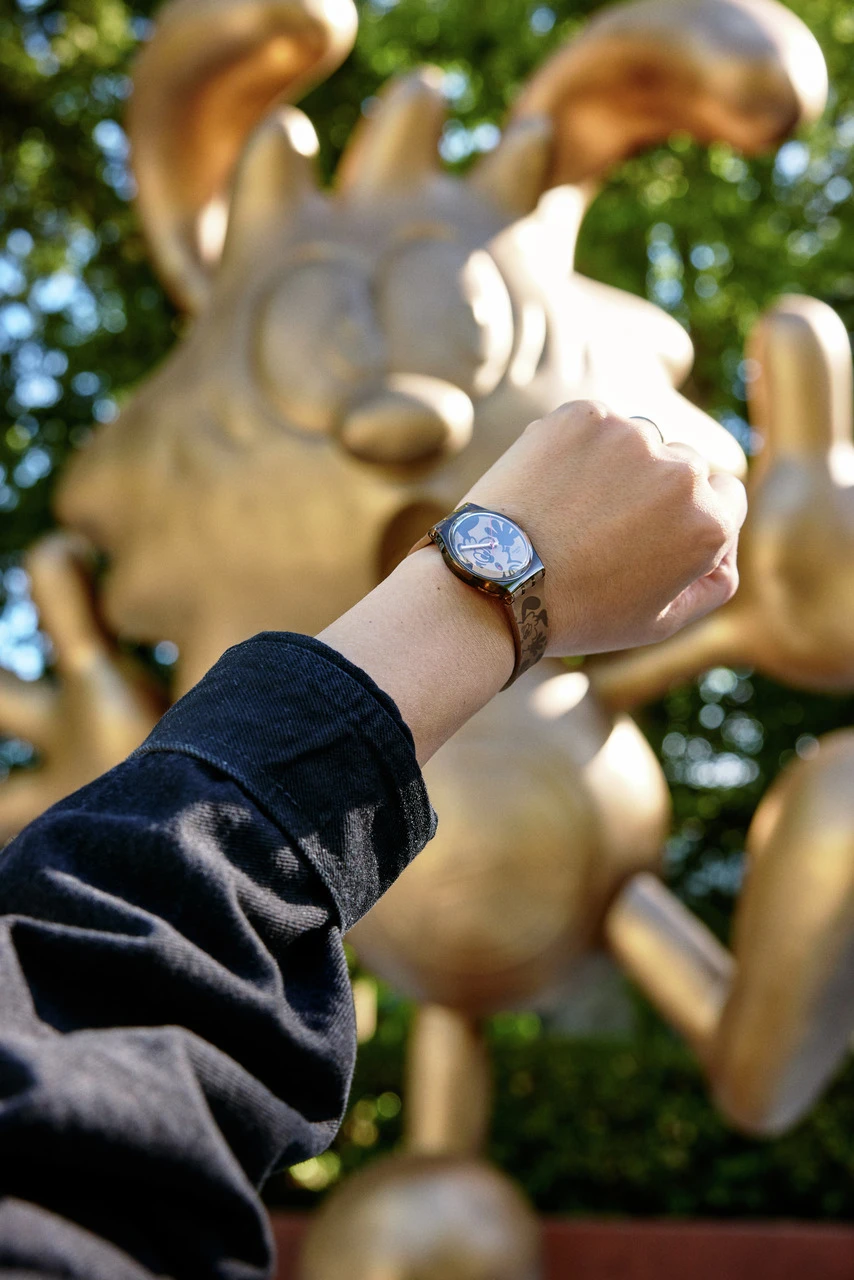 A person's wrist with a watch in the foreground, positioned against a blurred background showcasing a large golden statue shaped like a playful cartoon rabbit.