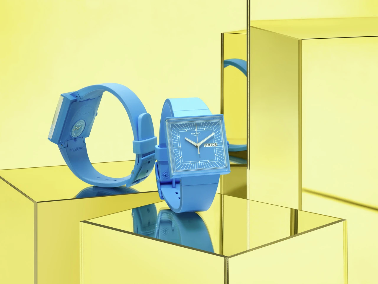 Two blue swatch watches displayed on reflective glass surfaces inside a yellow mirrored box setup, highlighting their unique square and round design details.