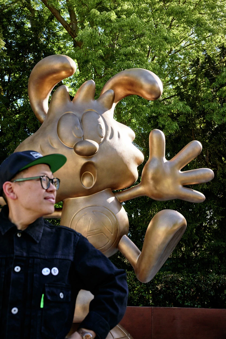 A young man wearing glasses and a snapback hat stands beside a large golden statue of a cartoon character with exaggerated features, poised in a playful stance. trees in the background.