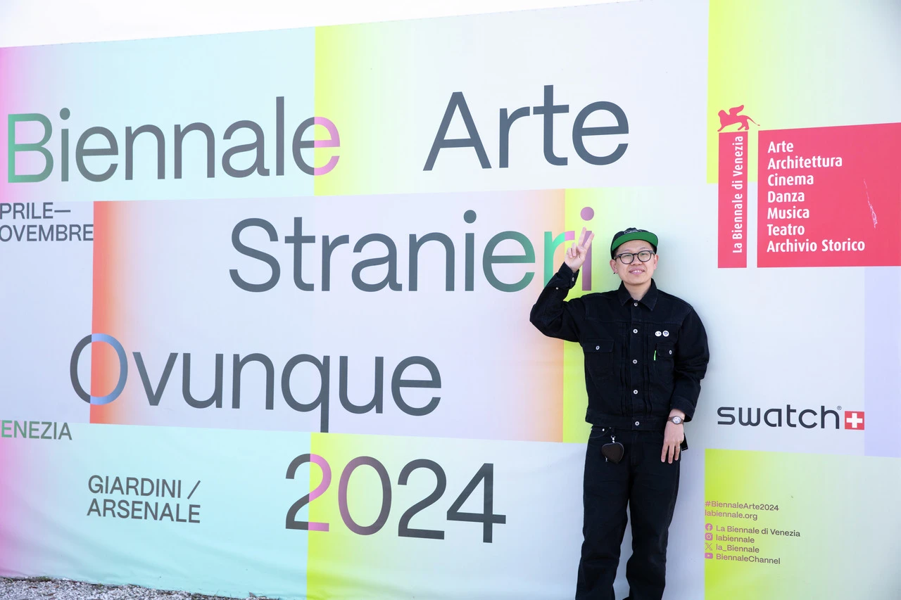 A person wearing a black outfit and cap salutes while standing in front of a colorful sign for the venice biennale 2024, titled "stranieri ovunque.