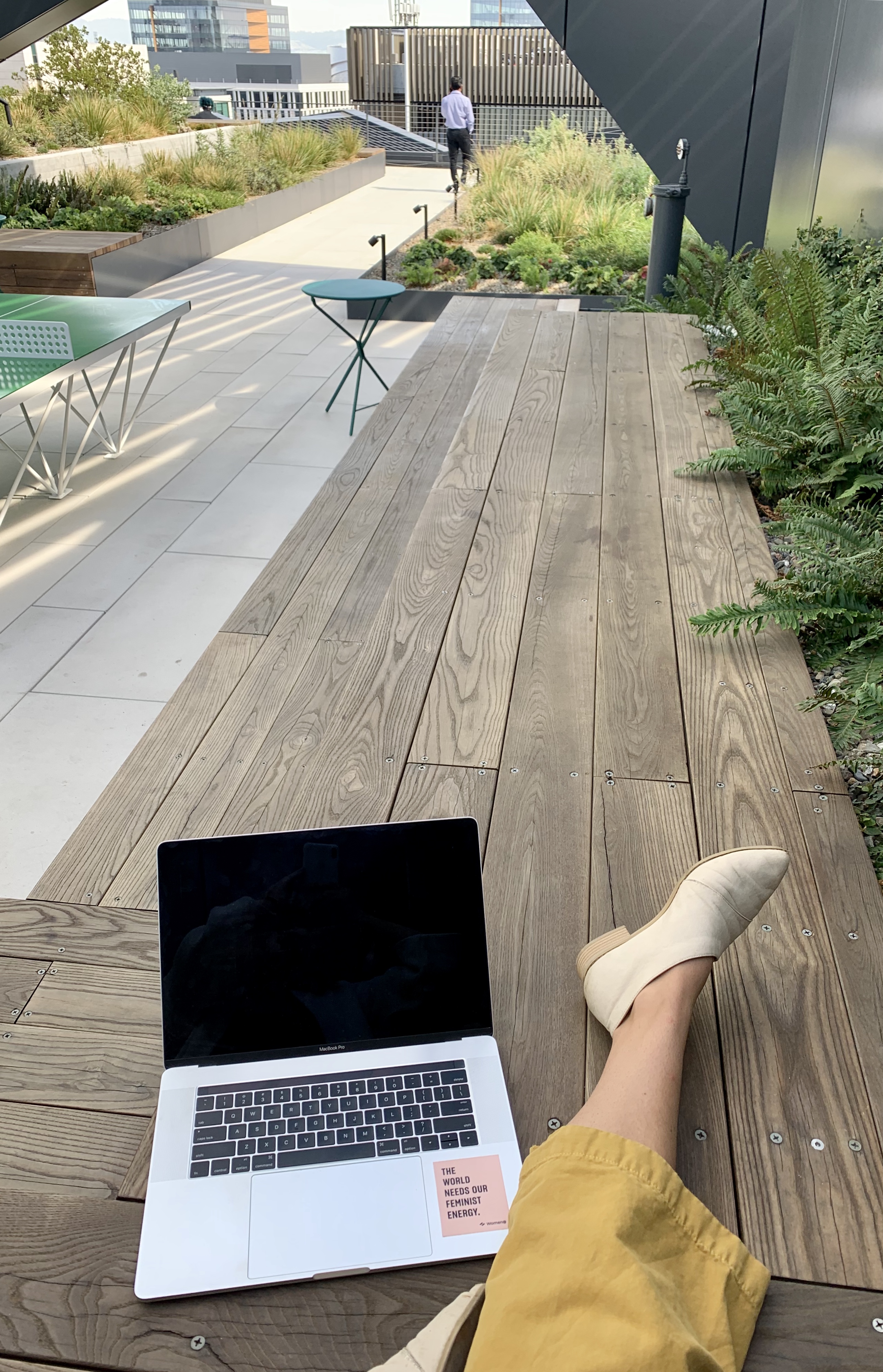 Emma sits on a bench with one foot out, computer resting on the bench. There is a laptop sticker that says "The world needs your feminist energy."