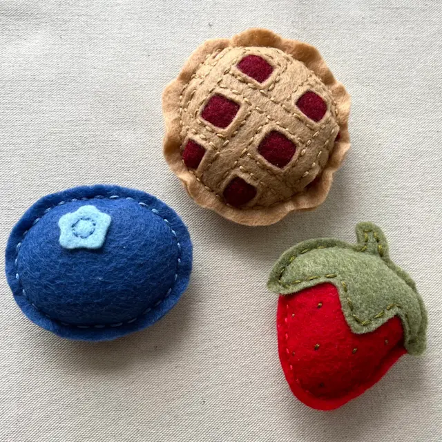 The image shows three small, hand-sewn felt toys filled with catnip that resemble miniature foods. Each item is made of soft, colorful felt and features simple stitching details:

The first item at the top is a small felt pie with a tan-colored crust and a lattice design on top, showing red felt underneath, resembling a berry pie. To the left, there’s a blueberry-like felt piece, made with dark blue felt and a small light-blue flower shape in the center, representing a blueberry or a blueberry tart. The third item, on the right, is a bright red felt strawberry with a green leafy top and small stitched details, giving it a textured appearance.
All items have a charming, handcrafted look and are arranged on a plain, light-colored fabric background.