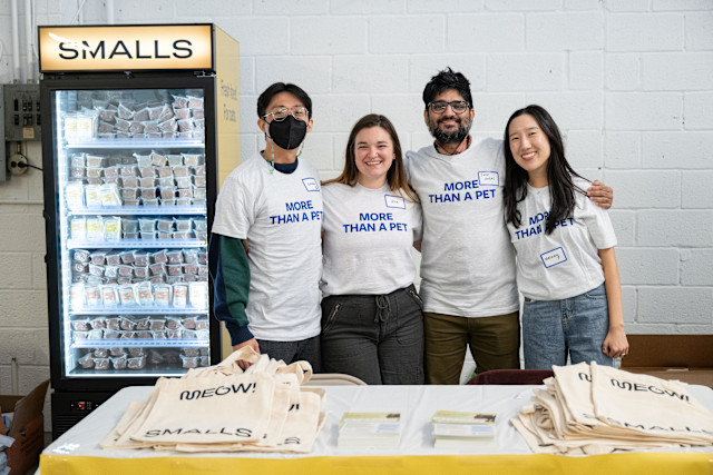 Smiling team members in branded 'More Than a Pet' t-shirts pose at a Smalls event. A refrigerator stocked with fresh cat food and tote bags reading 'Meow!' are visible, showcasing Smalls' dedication to premium feline nutrition.