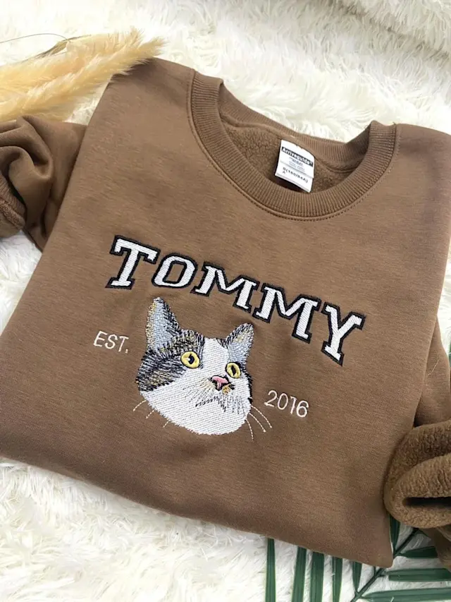 Cozy embroidered sweatshirt featuring a cute tabby cat design and 'TOMMY EST. 2016' text. Perfect for cat lovers, this warm, brown fleece-lined pullover combines comfort and style for casual wear.