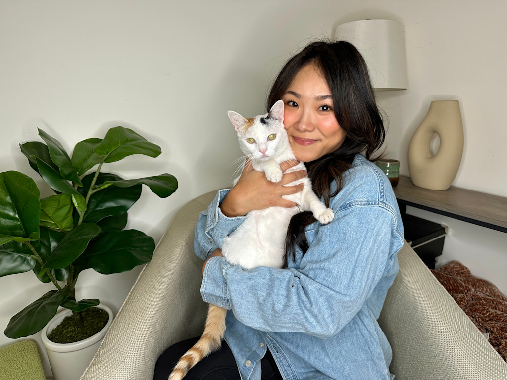 Grace Choi, a woman with long, dark hair is sitting on a light-colored armchair, holding a white cat with patches of orange and black on its head and tail. The woman is smiling warmly at the camera, wearing a light blue denim jacket. The cat appears calm and relaxed in her arms. To the left of the woman, there is a large, green potted plant with broad leaves, adding a touch of nature to the setting. Behind the woman on the right side, there is a wooden shelf with a beige, abstract-shaped vase and a green ceramic container. The background is simple and neutral, allowing the focus to remain on the woman and her cat.