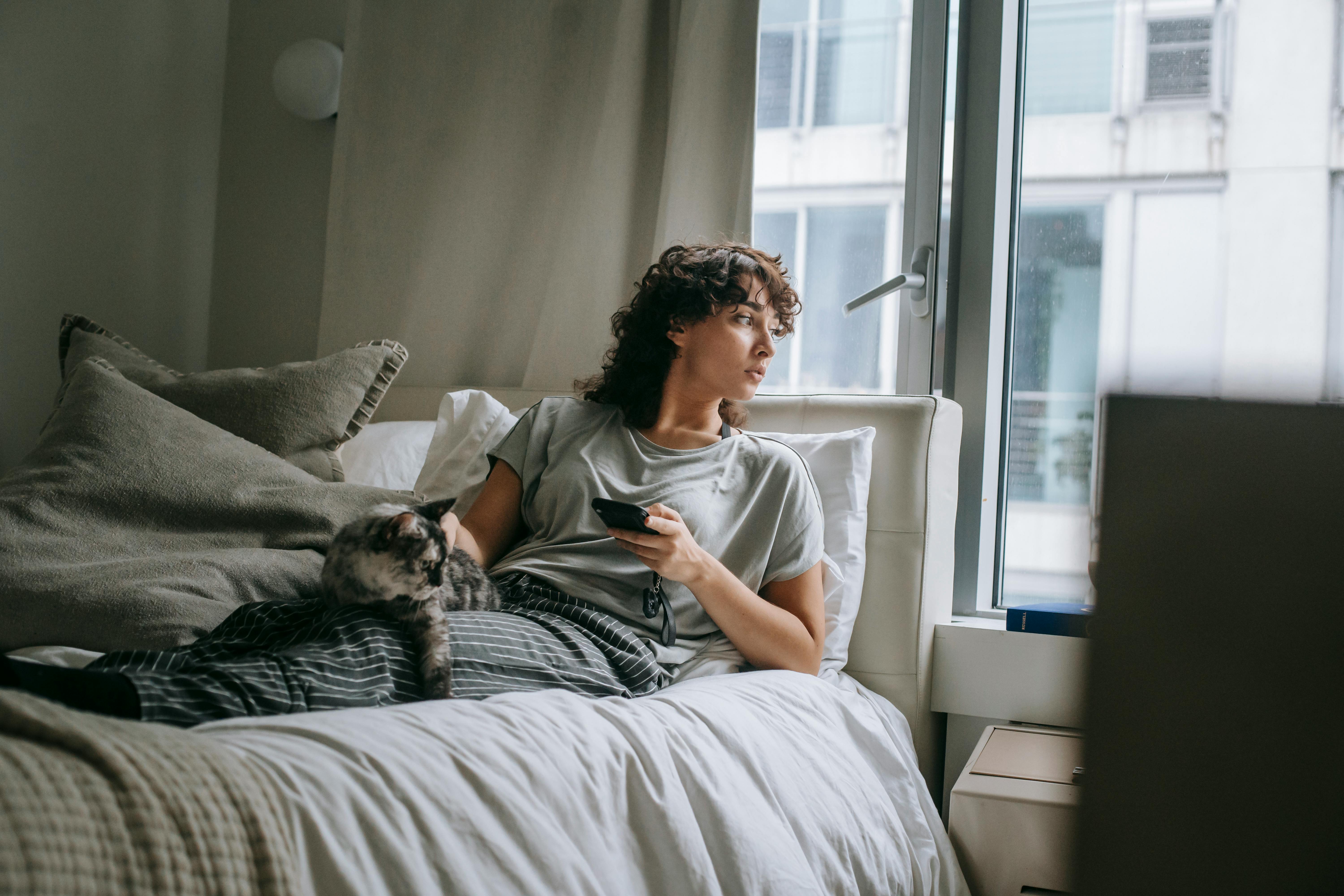 Calm woman sitting on bed with both legs stretched out, her grey-black marbled cat laying on them. While glancing out of the window, she has one hand petting her cat and the other holding her smart phone.