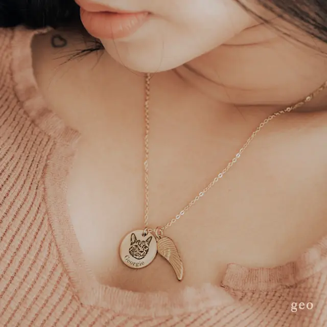 The image shows a close-up of a woman wearing a delicate necklace with two charms. One charm is a round pendant featuring an engraved illustration of a cat’s face, which has pointed ears and a gentle expression. The other charm is a small, wing-shaped pendant, adding a whimsical touch to the necklace. 