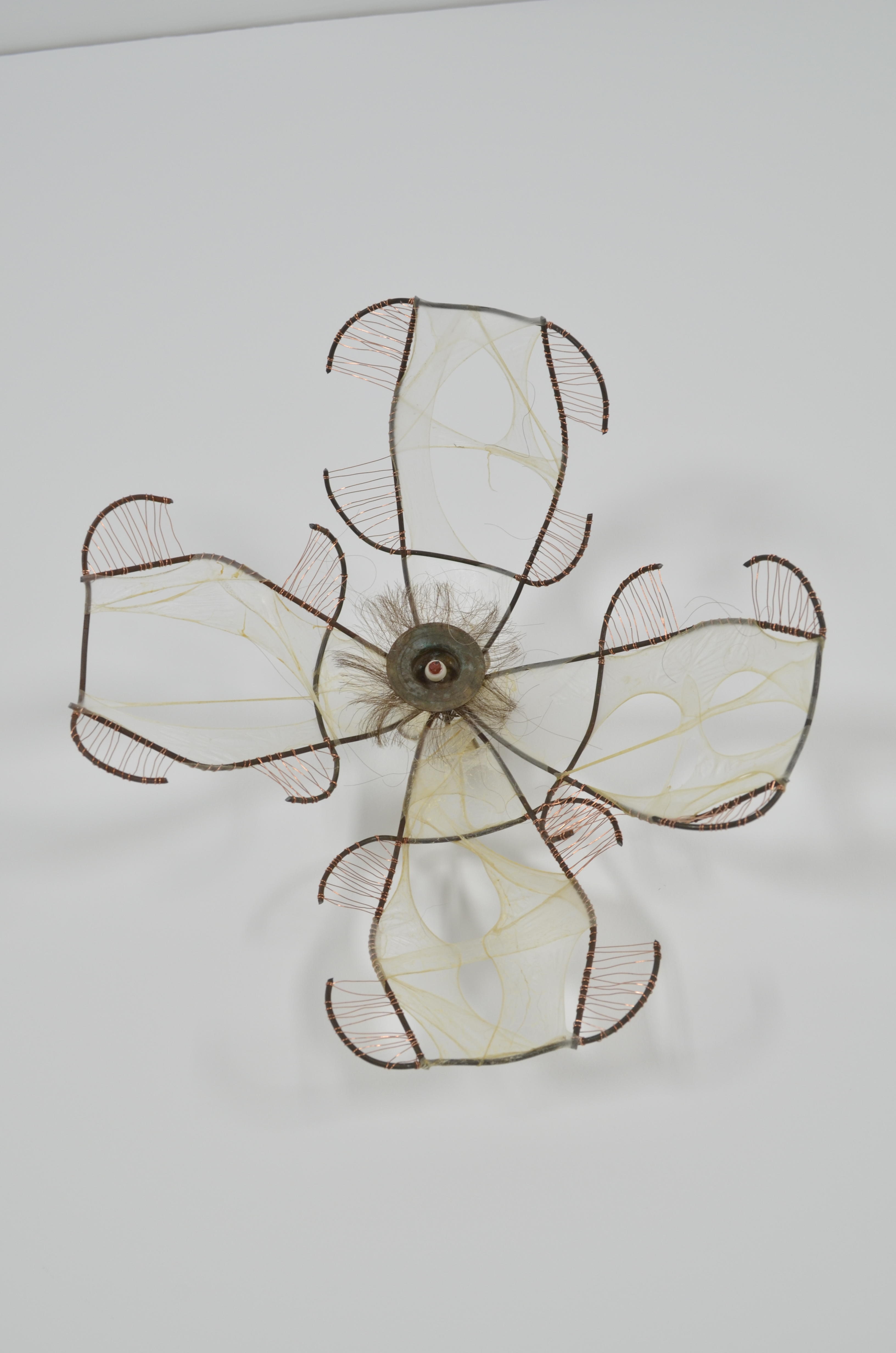 This dogwood flower pins seamlessly into the wall.  It is an assemblage of found items (bead, round metal item, wire fence), wire, gut, & hair.