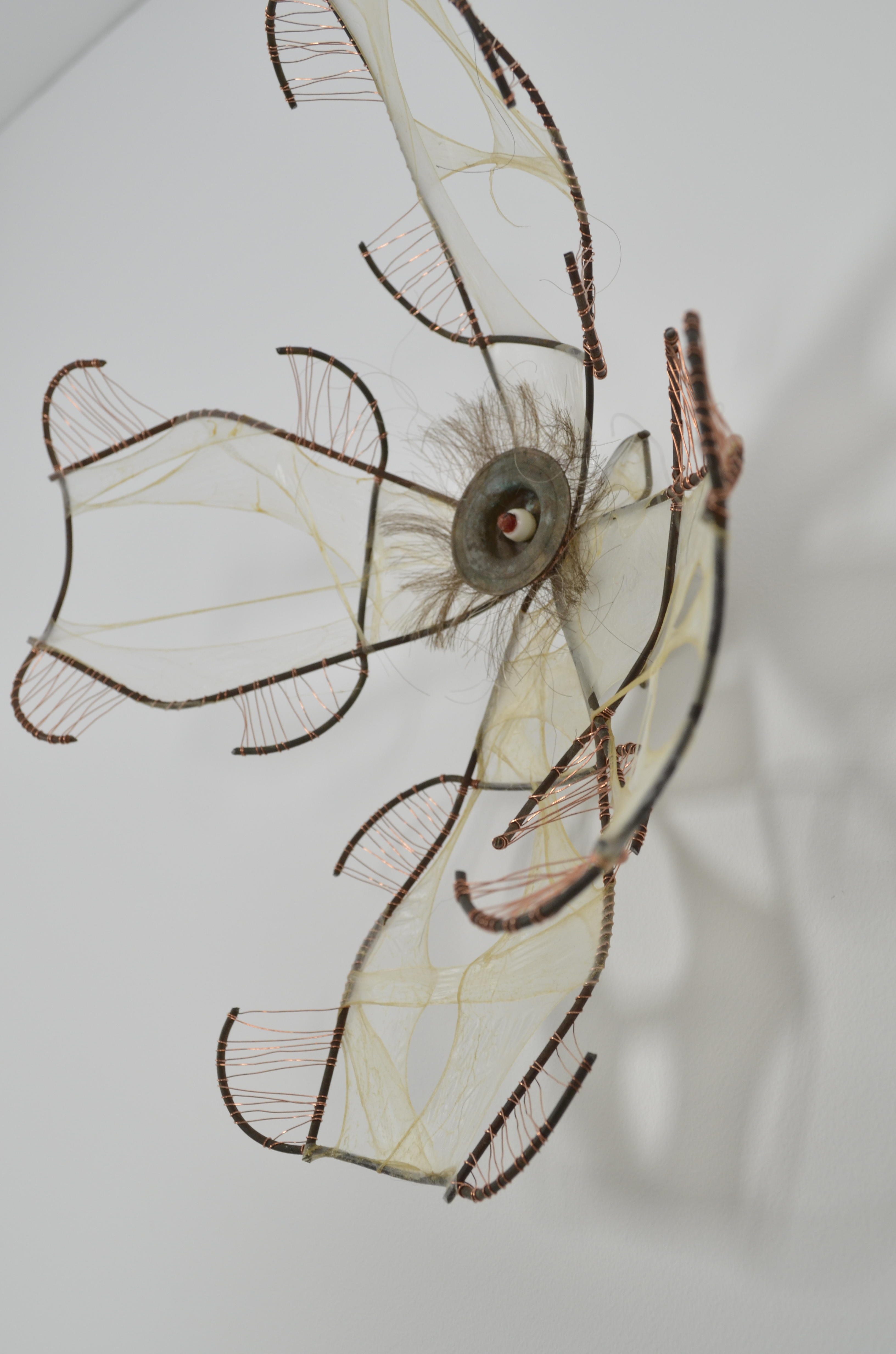 This dogwood flower pins seamlessly into the wall.  It is an assemblage of found items (bead, round metal item, wire fence), wire, gut, & hair.
