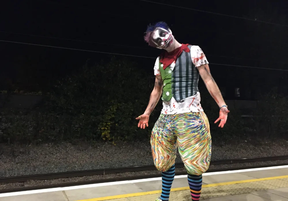 Charlie Jackson dressed as a clown holding his arms out to the sides in a powerful pose whilst stood on a train platform