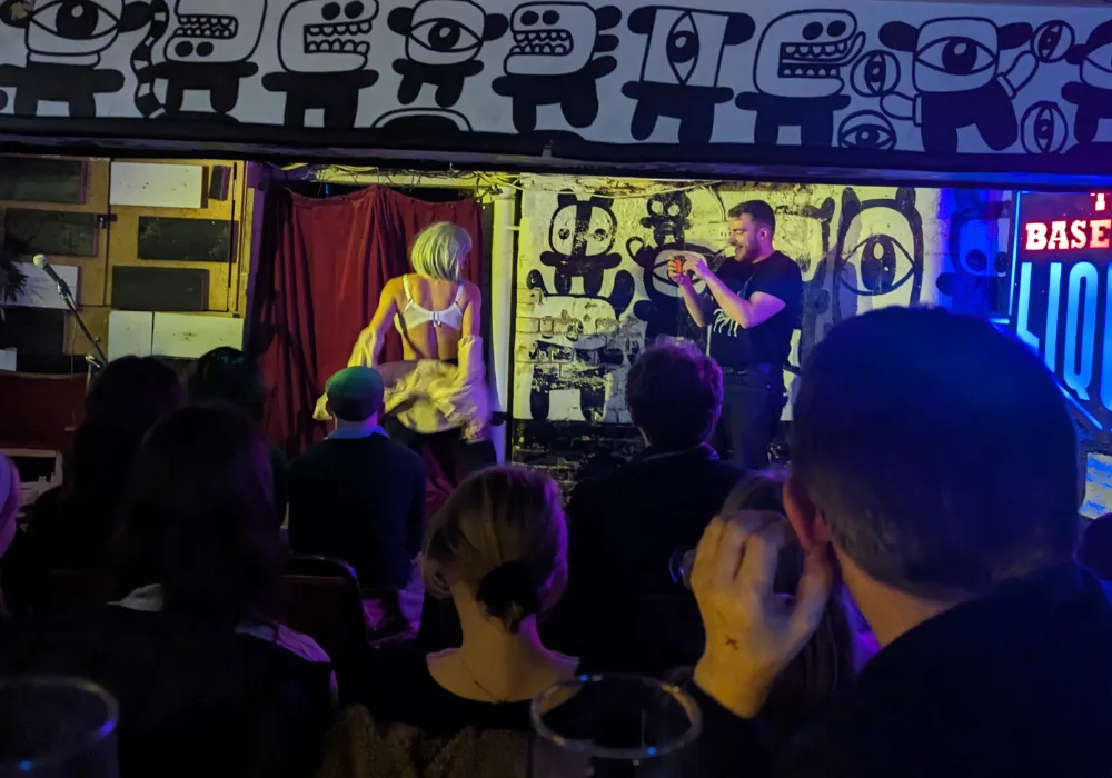 Comedian on stage dressed as an old lady, taking her shirt off to reveal a white bra as an audience member takes a photo on his phone