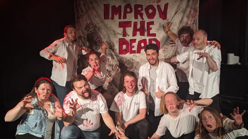 The Improv the Dead comedy troupe. 10 Improvisers with bloodied tops in front of a sign reading "IMPROV THE DEAD"