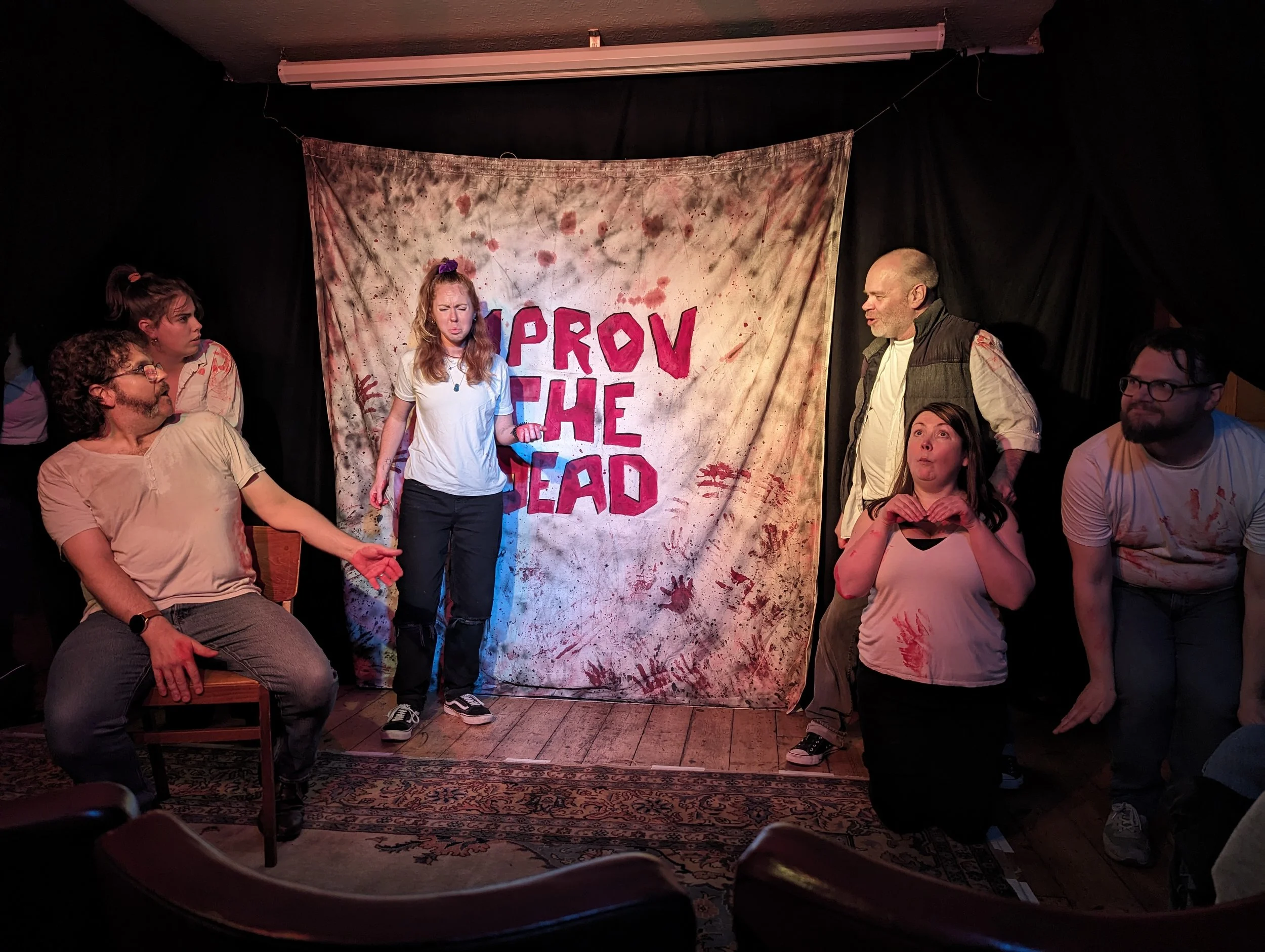 Improv the dead on stage. Three cast members are kneeling like otters, one is sat down and pointing whilst another looks confused.