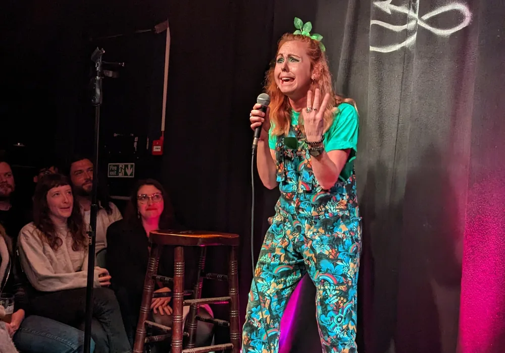 Viki Jackson on stage performing comedy holding a microphone with a pained expression on her face