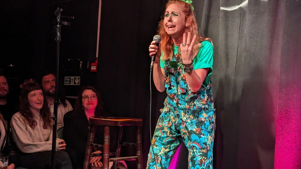 Viki-standup Viki Jackson on stage performing comedy holding a microphone with a pained expression on her face