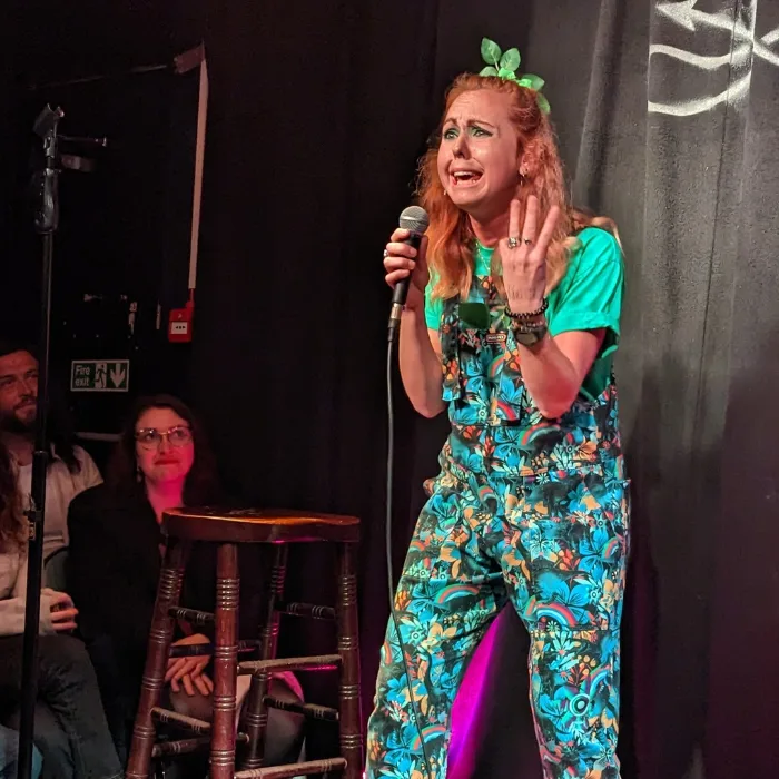 Viki-standup Viki Jackson on stage performing comedy holding a microphone with a pained expression on her face