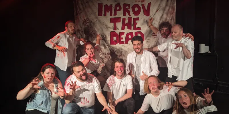 Improv team (10 people) posing like zombies in front of blood covered sign saying improv the dead 