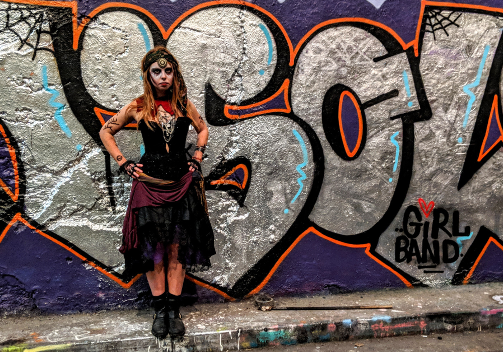Viki Jackson dressed as a fortune teller with face paint stood with her hands on her hips besides some graffiti