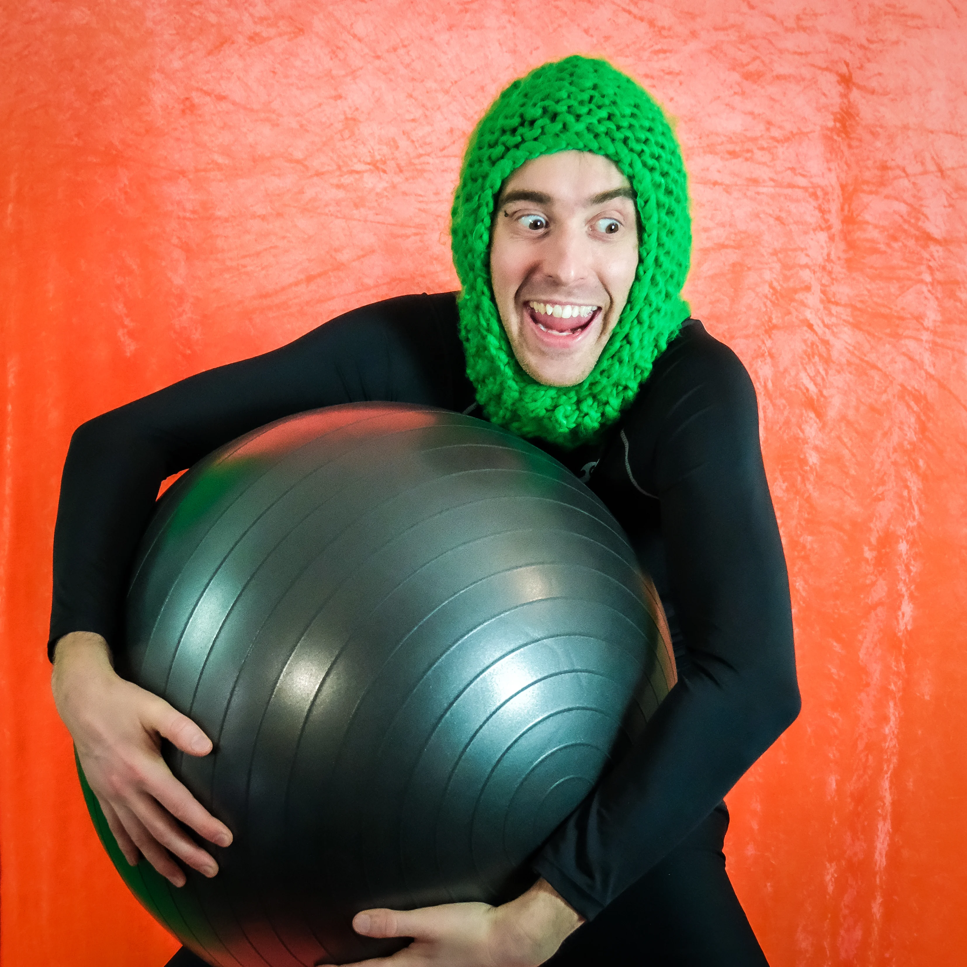 Charlie Jackson holding an exercise ball and wearing a knitted green hood. Otherwise he's dressed all in tight black. There's an orange background. He kinda looks like a goblin.
