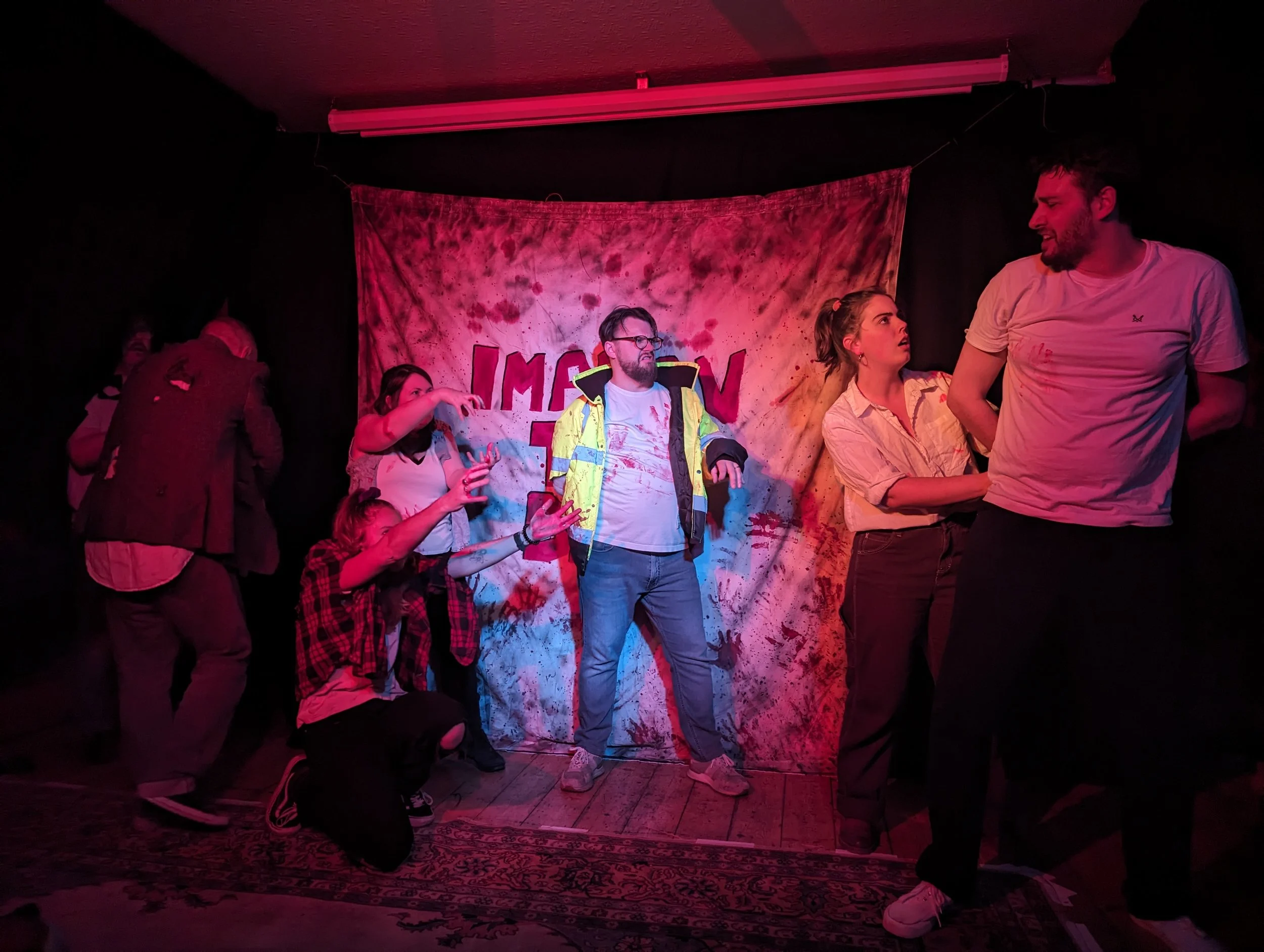 Improv The Dead team on stage. Two team members have their arms outstretched trying to reach another person who looks shocked meanwhile another member is arresting another.