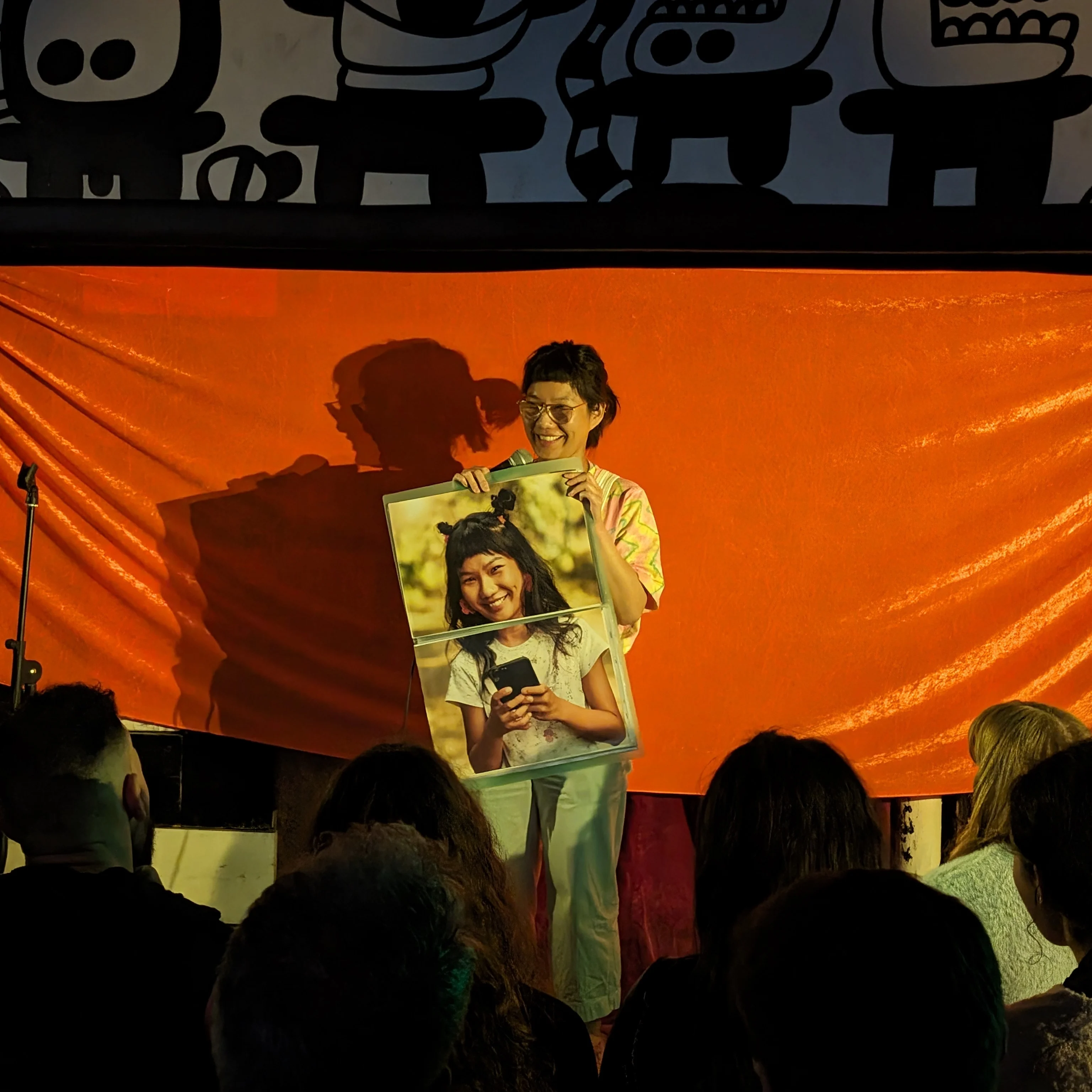 Jen Zheng Comedian on stage holding a microphone and a photo of themselves