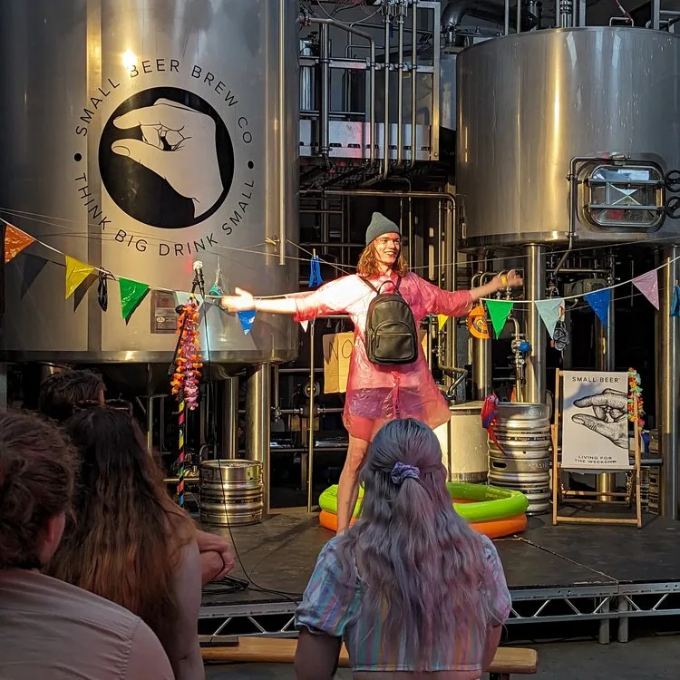modern art variety July Cam Comedian on stage wearing a pink waterproof and a backpack on their front