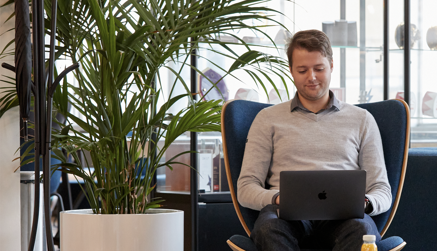 A photo of an office worker sitting in an armchair in the co-working area of the modern office, typing on his laptop.