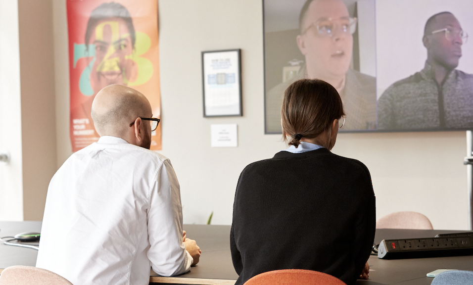 A photo of a hybrid office meeting. Two coworkers are in the meeting room at the table while the other two are on a large screen joining remotely.