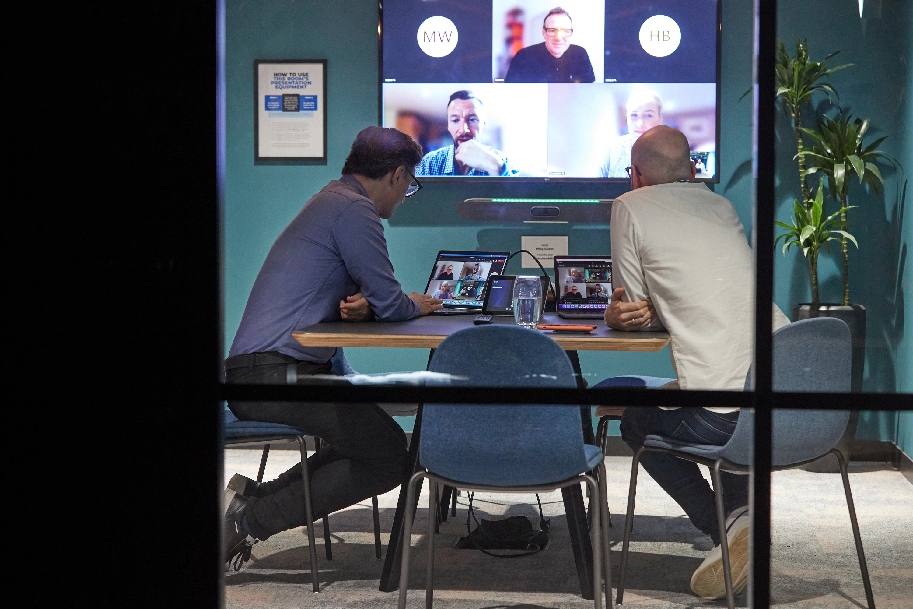 A photo of five co-workers in a meeting room. Tow of them are in the room, while three are on the large screen on the wall, joining remotely.