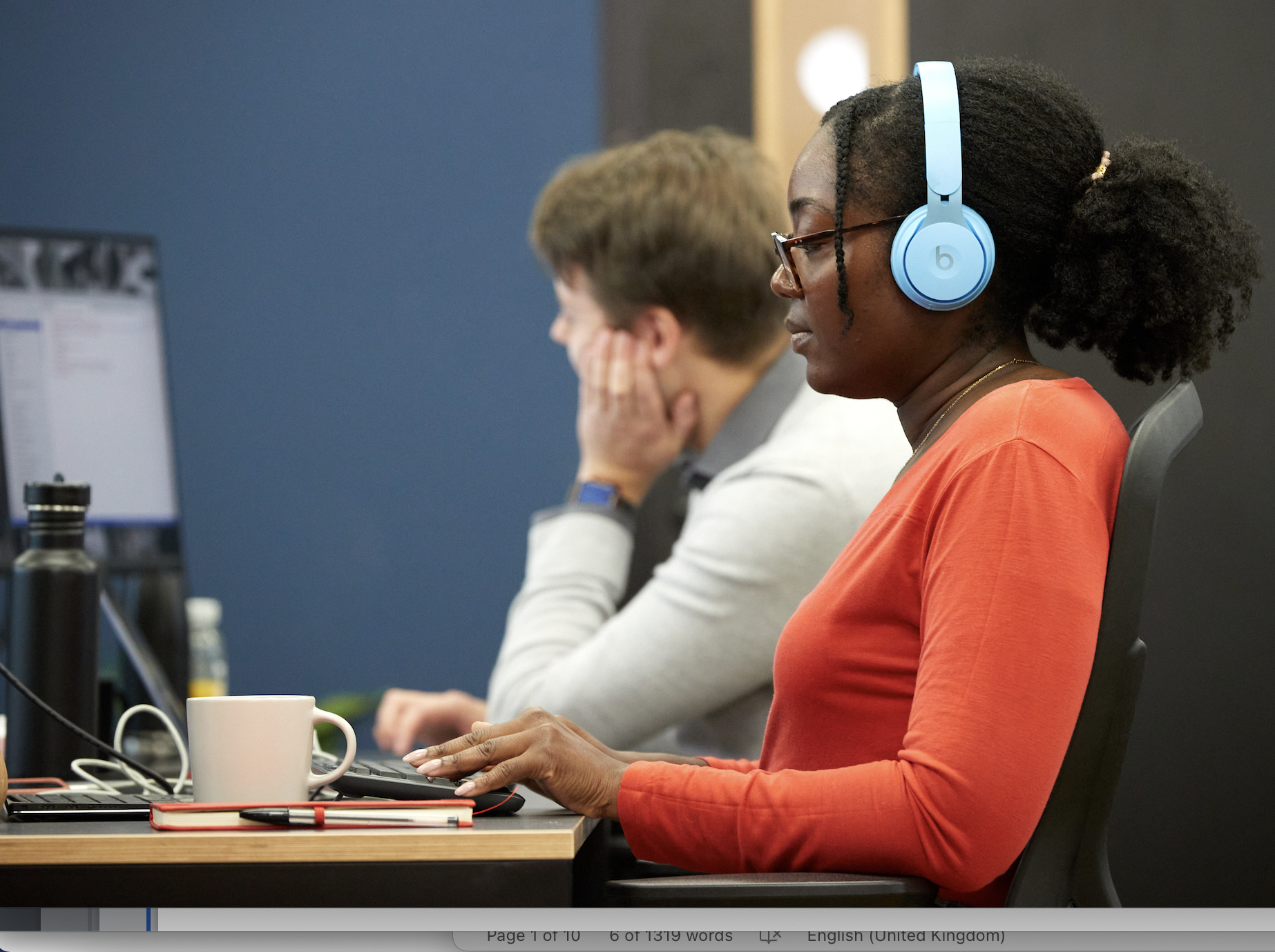 A photo of a woman working on her laptop at the desk, wearing bright blue headphones.